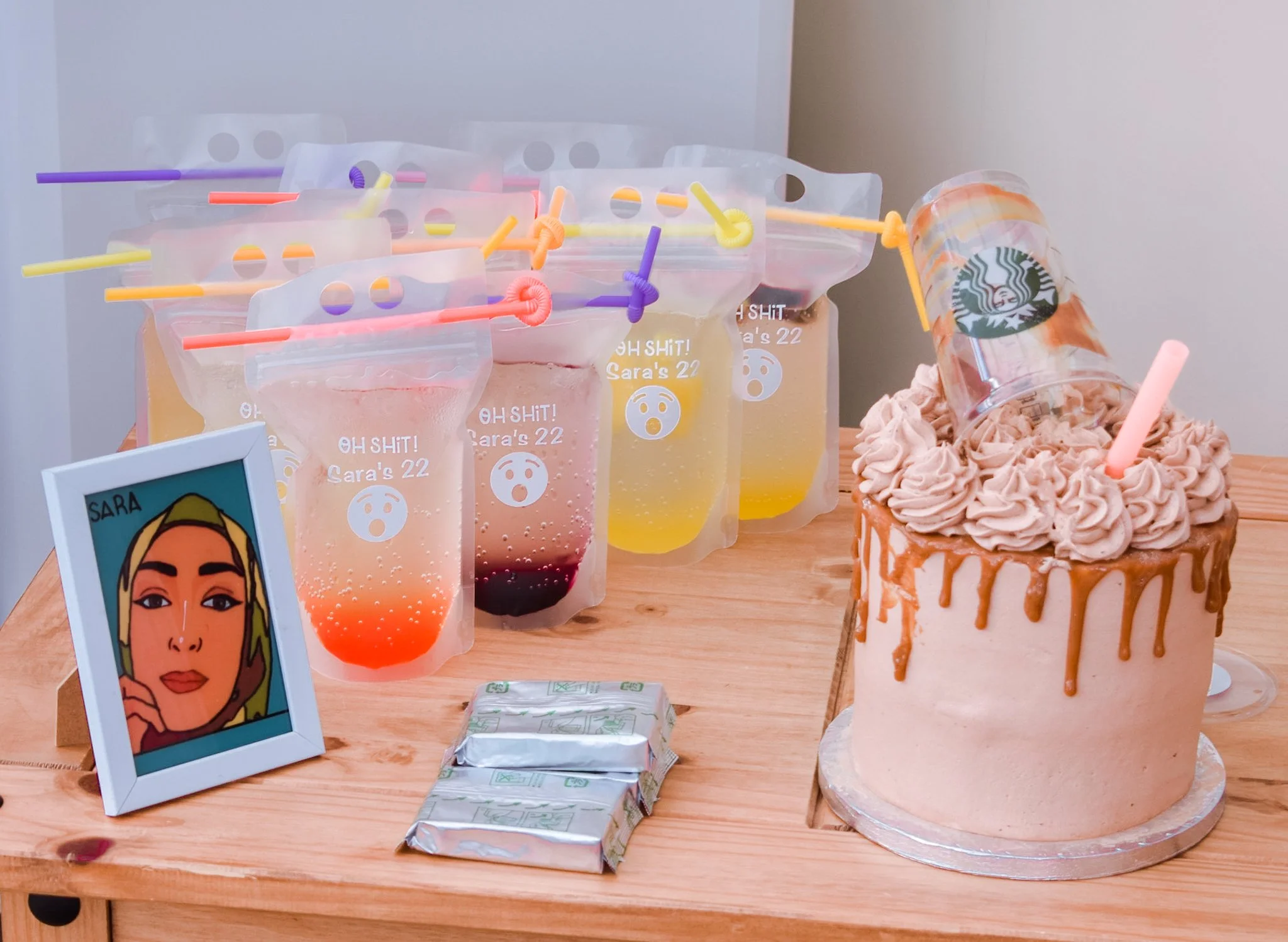 A birthday table with a cake topped with frosting and a pink straw, several colorful drinks in plastic bags with straws, a framed drawing of a woman, and wrapped gifts.