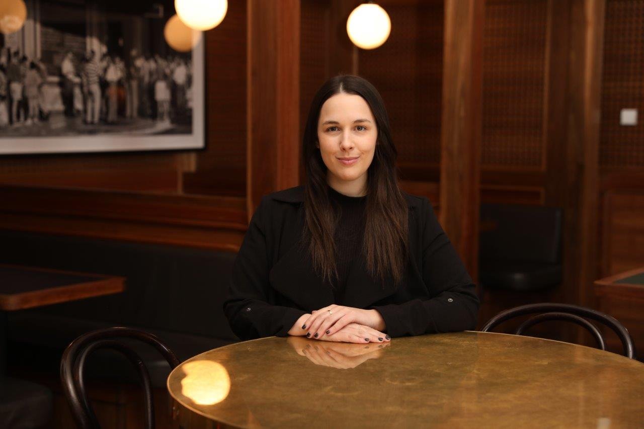 A woman with long dark hair, dressed in black, sitting at a round wooden table in a dimly lit restaurant or cafe with wooden decor and hanging round lights.