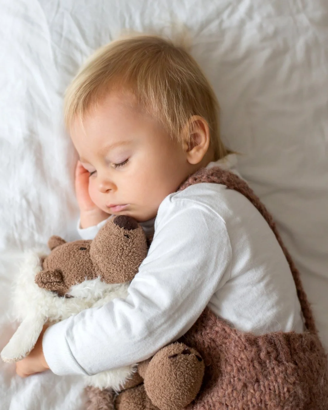 A young child sleeping peacefully on a white pillow, hugging a stuffed teddy bear.