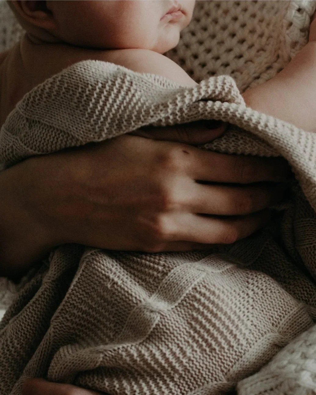 Close-up of a parent's hand holding a sleeping baby's torso, with the baby wrapped in a beige and brown knit blanket.