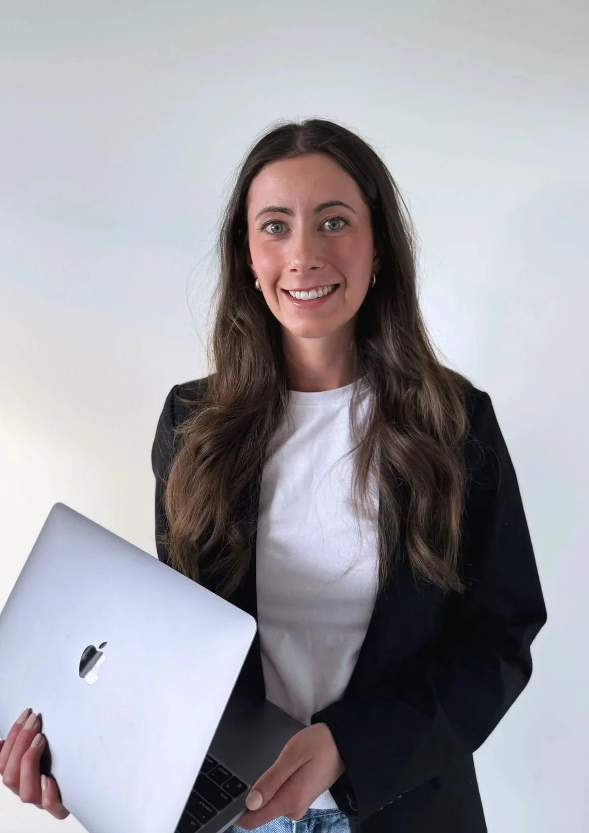 A woman with long brown hair, wearing a black blazer and white shirt, holding a silver MacBook laptop, standing against a plain white background, smiling at the camera.