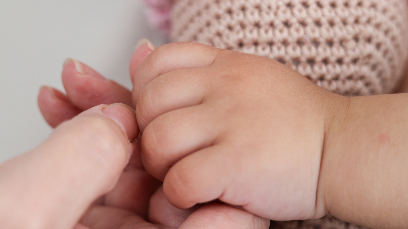 Close-up of an adult holding a child's hand, with a background of pink knitted fabric.