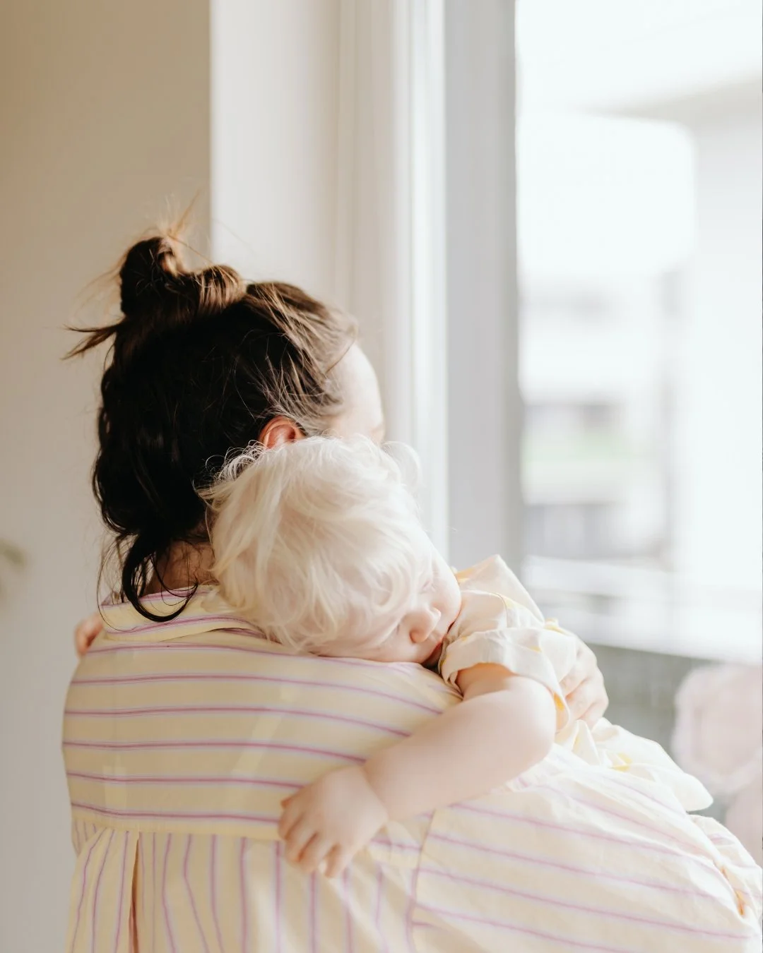 A woman holding a sleeping child by a window in a bright room.