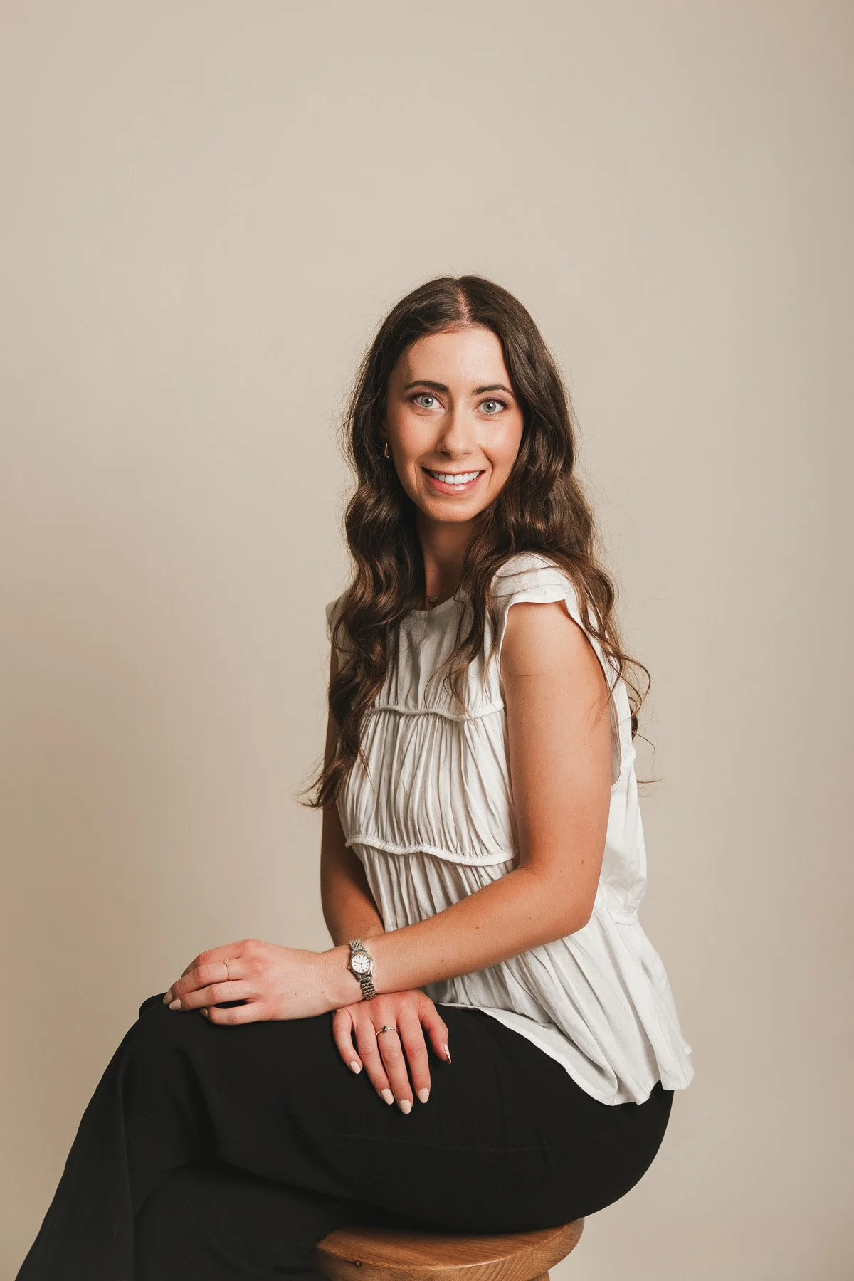 A woman with long wavy brown hair, wearing a white ruffled top and black pants, sitting on a wooden stool against a plain light-colored background, smiling and looking at the camera.