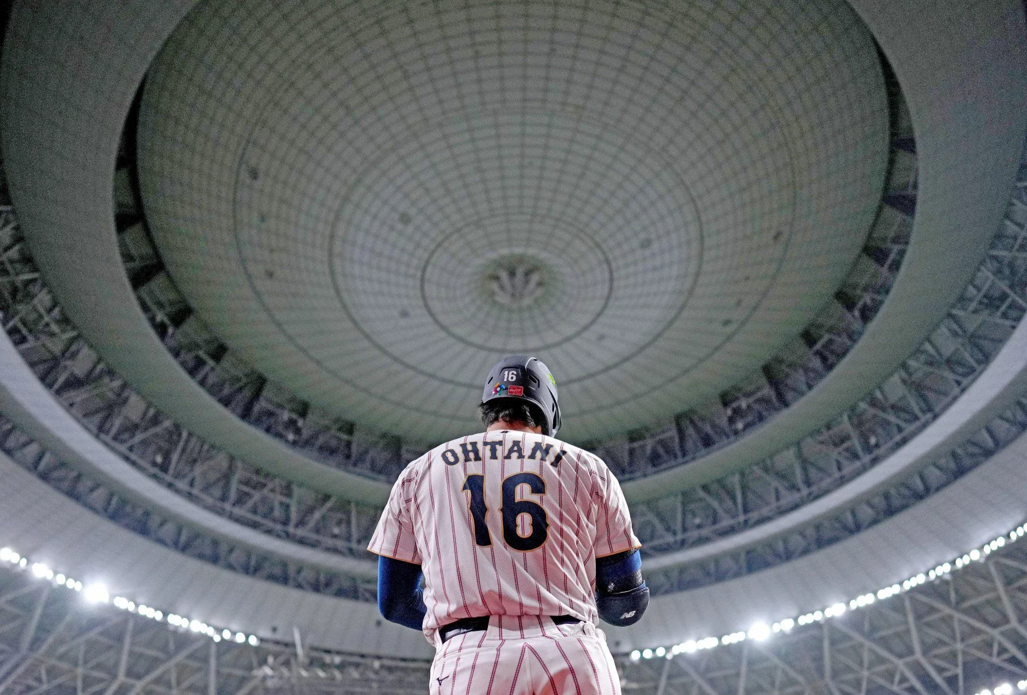 Baseball player standing in the center of a large stadium with a domed ceiling, viewed from behind. The player is wearing a white uniform with red pinstripes, the number 16, and the name 'OHTANI' on the back, along with a helmet.