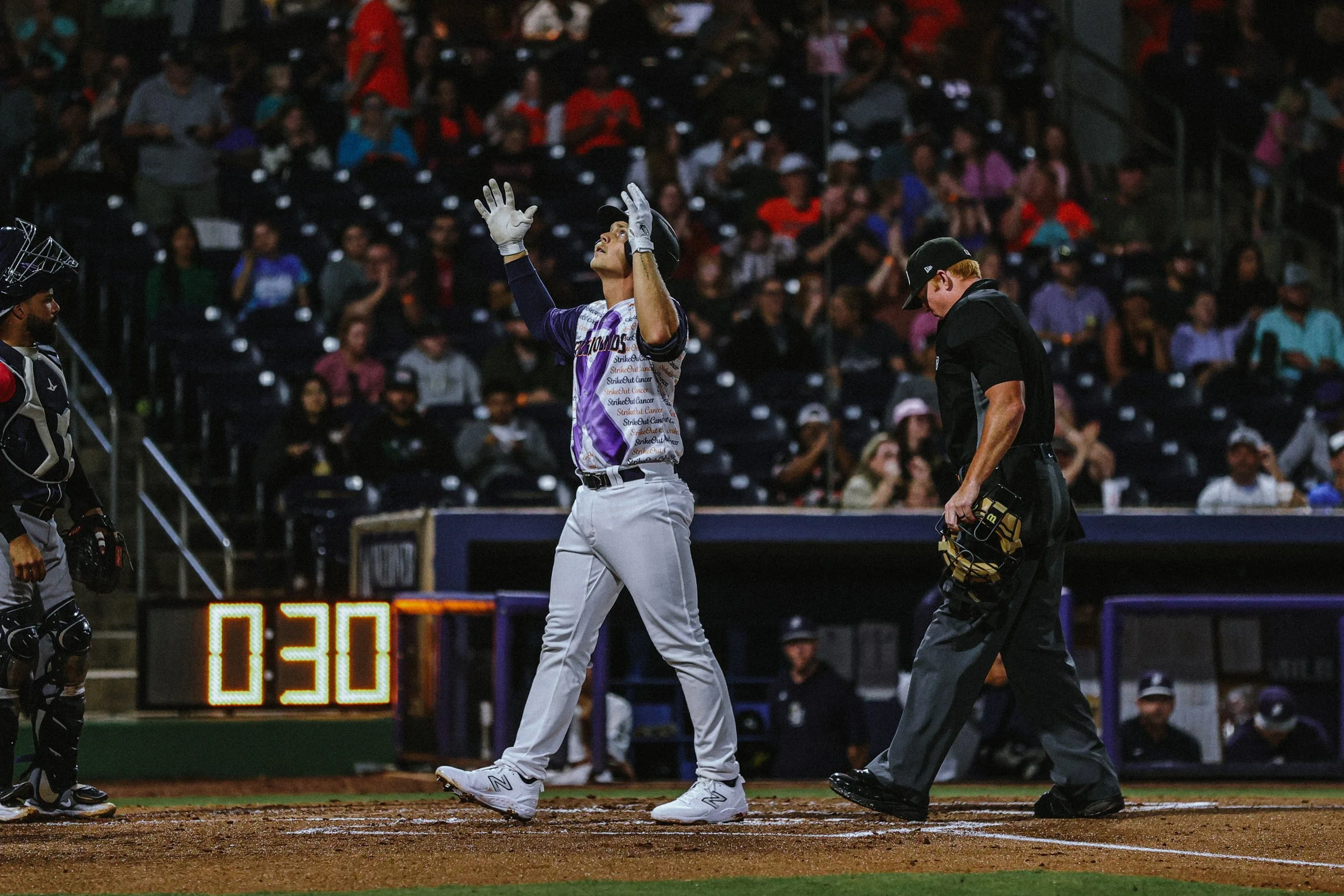 A baseball player in purple and gray uniform celebrates with raised hands at home plate. An umpire is nearby looking down. A catcher is visible to the left and a scoreboard shows 0:30 in the background, with a crowd of spectators in the stands.