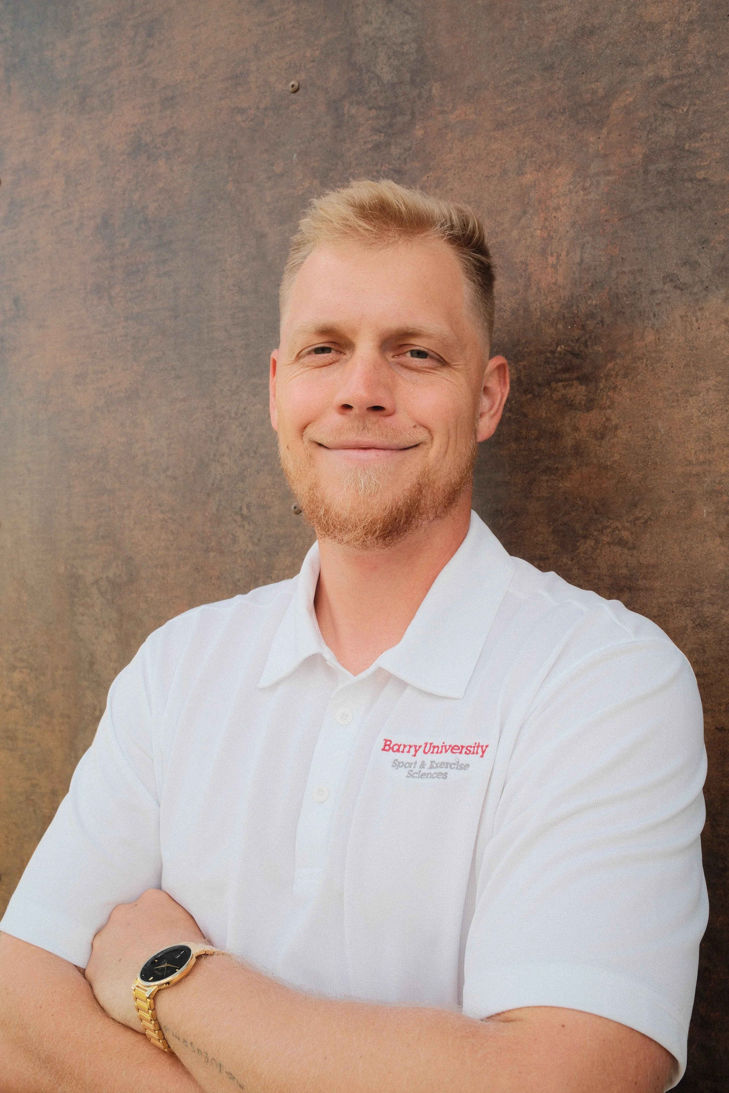 A smiling man with short blond hair and a beard, wearing a white polo shirt with a Barry University logo, standing with arms crossed in front of a textured brown wall.