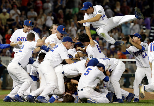 Baseball players in team uniforms celebrating on the field after victory, some jumping and others bending down, with spectators in the background.