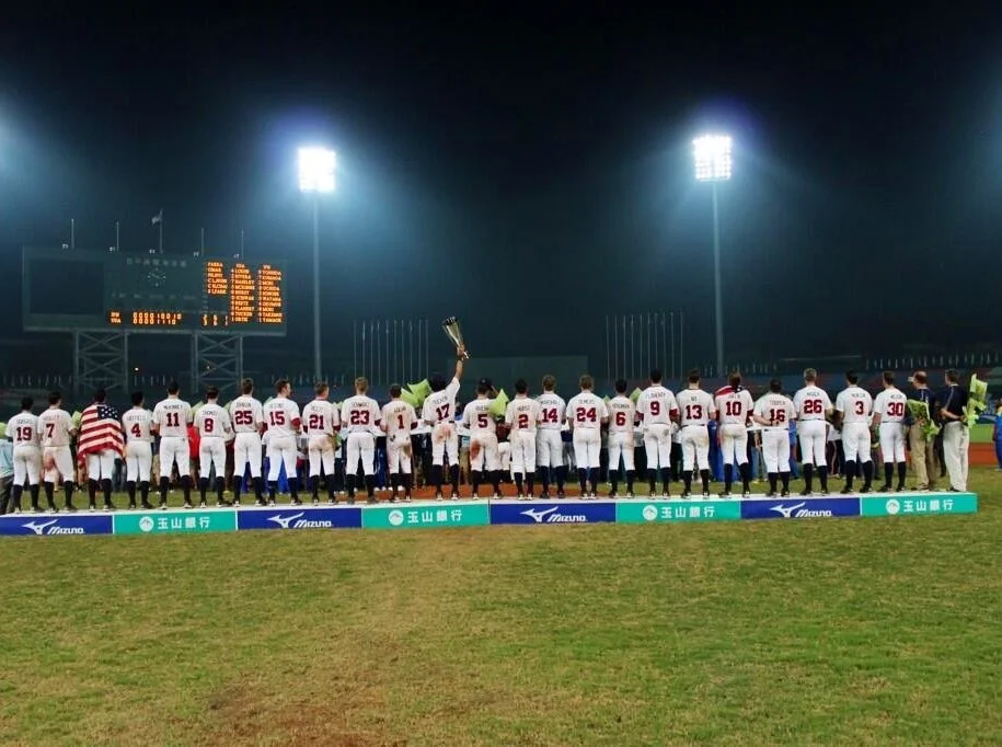 Baseball team standing on field at night in a row during a ceremony, some holding a trophy, with game scoreboard and bright stadium lights in the background.