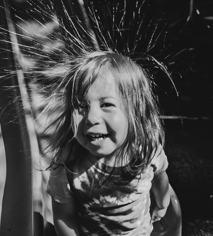 A young girl with PWS, long hair, smiling and looking at the camera while playing outdoors in black and white.