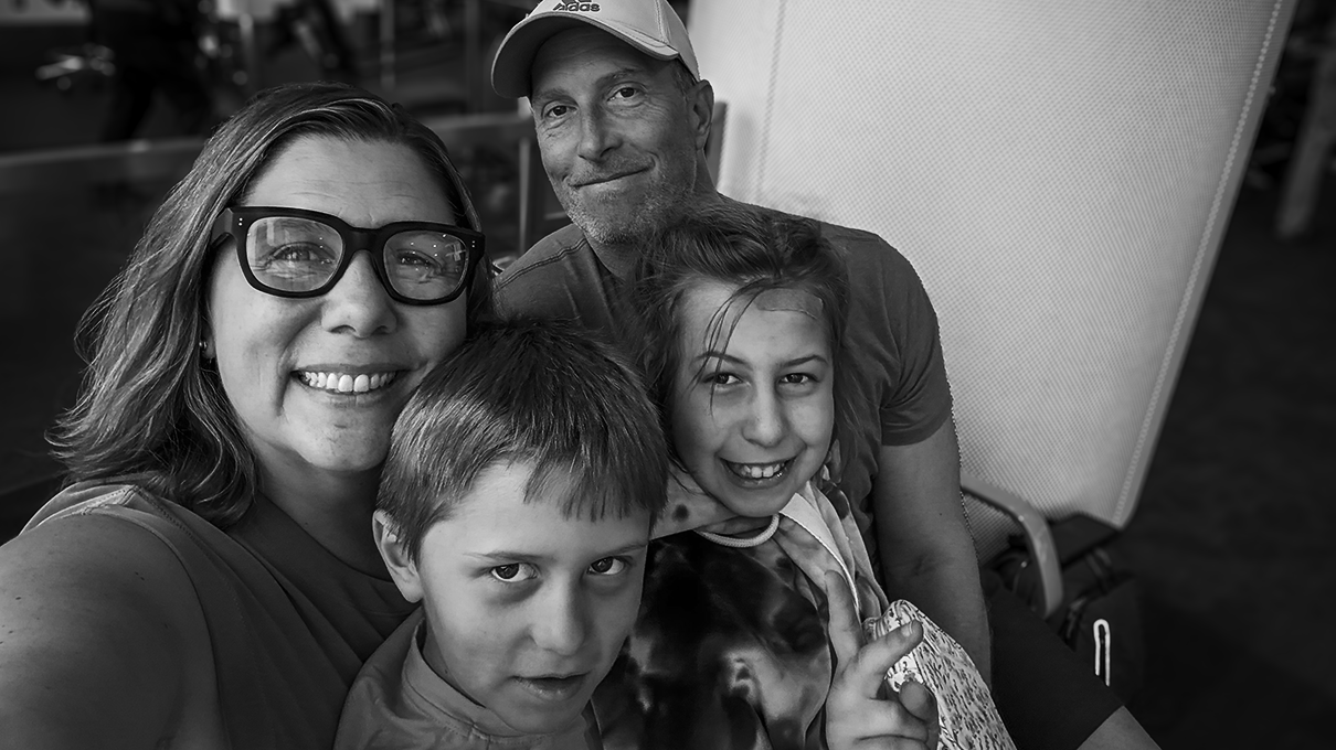 A family of four with a woman wearing glasses, a man with a cap, and two children, a boy and a girl, sitting closely together and smiling at the camera.