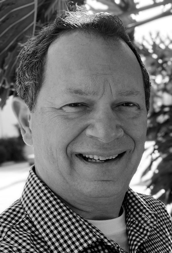 Black and white close-up photo of a man smiling outdoors, wearing a checkered shirt.