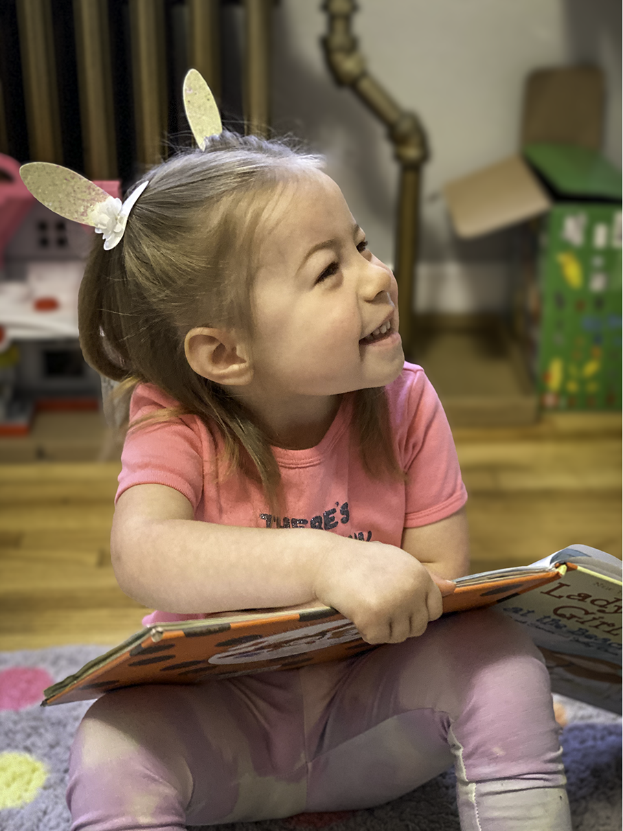 A young girl with PWS wearing bunny ears headband, a pink shirt and pink pants, sitting on the floor and reading a book, smiling and looking to the side.