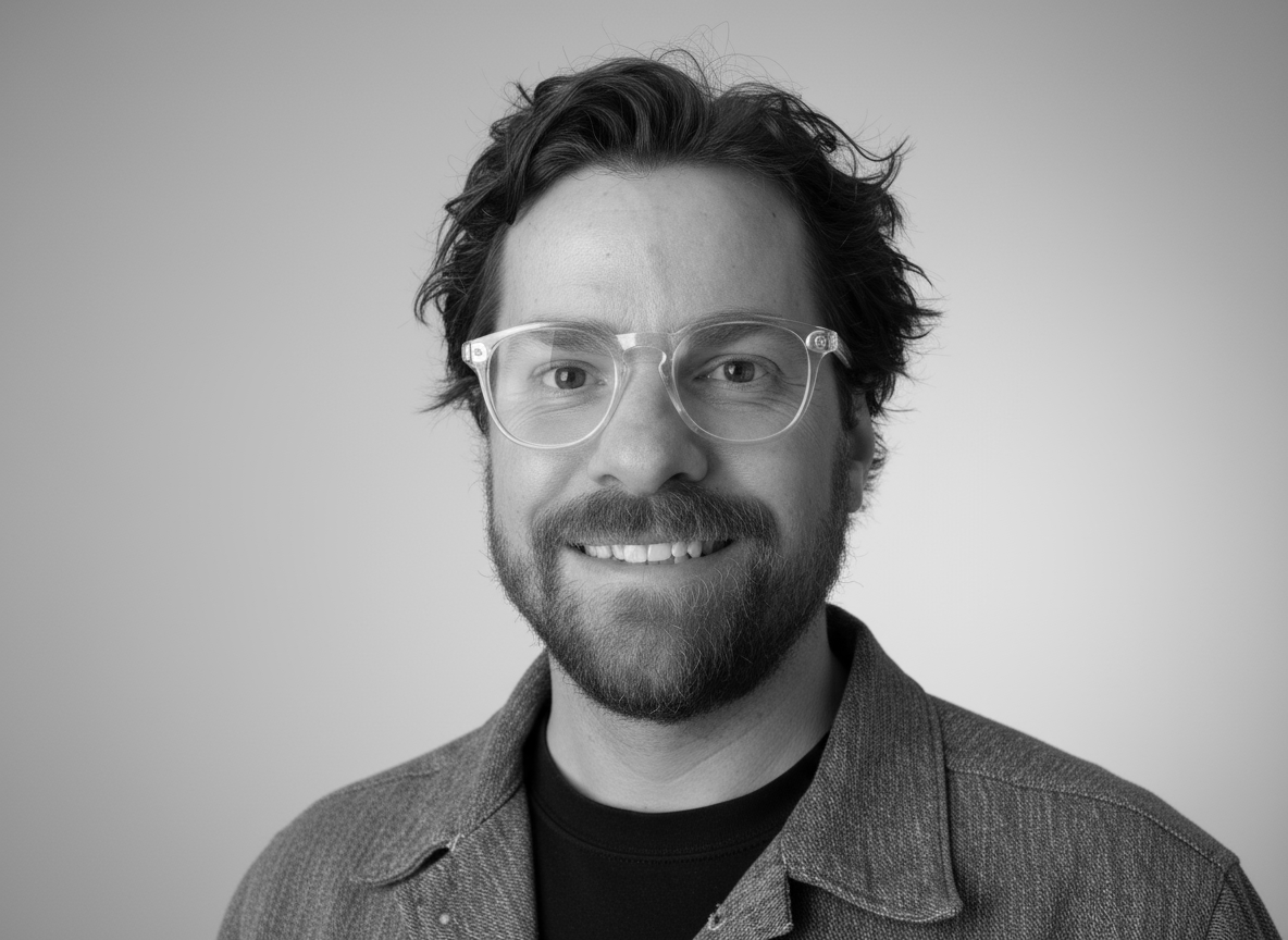Black and white portrait of a man with wavy hair, glasses, beard, and mustache, smiling, wearing a jacket over a shirt, against a plain background.