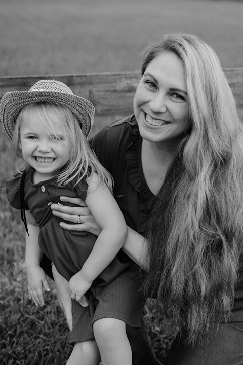 A woman with long hair and a young girl wearing a hat smiling outdoors.