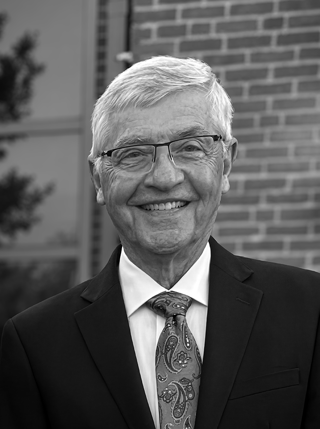 An man with glasses and a smile, dressed in a suit and tie, standing outdoors in front of a brick building.
