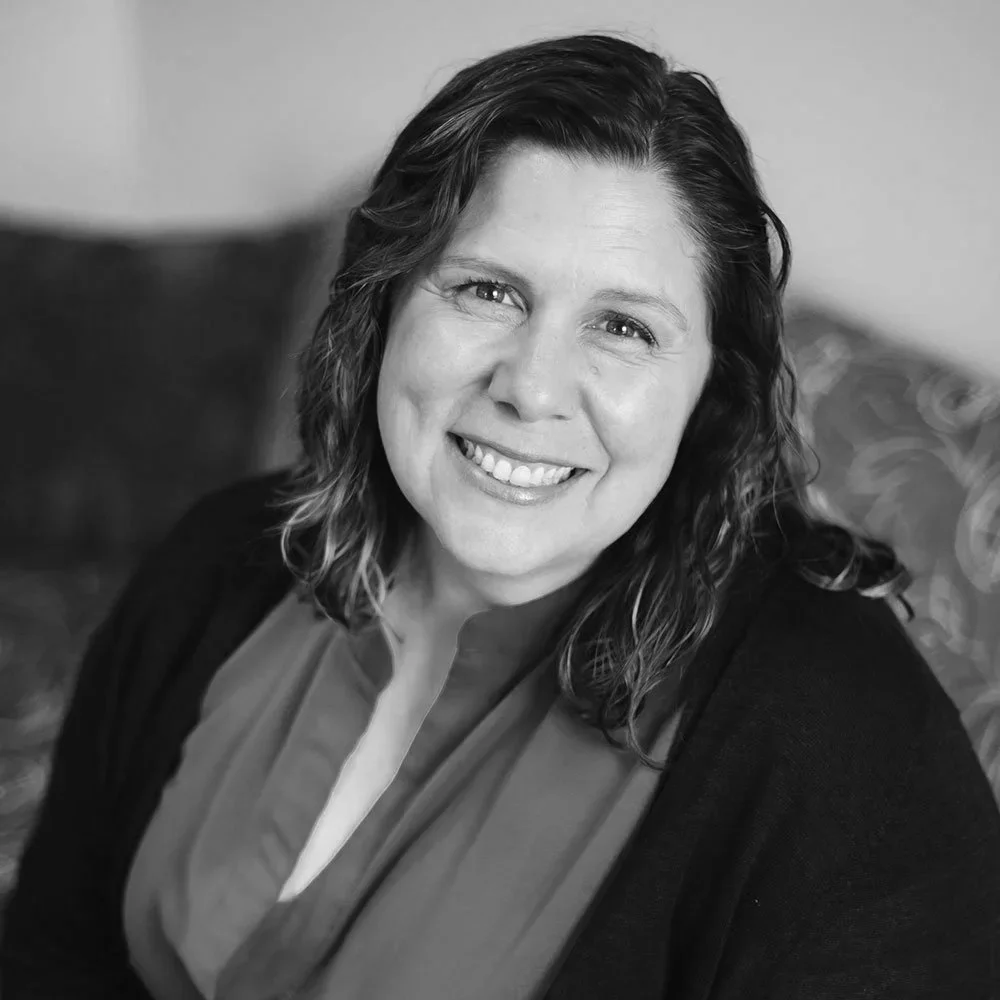 Black and white portrait of a smiling woman with shoulder-length wavy hair, wearing a scarf over a blouse, sitting in front of an out-of-focus background.