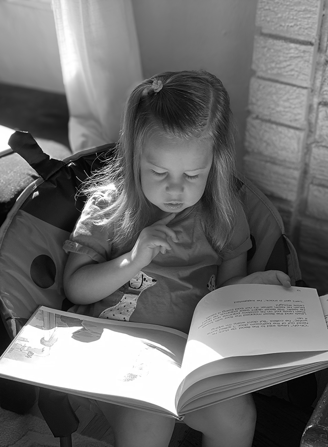A young girl with long hair, sitting, is looking at an open book with sunlight illuminating the pages. She appears focused on the book.