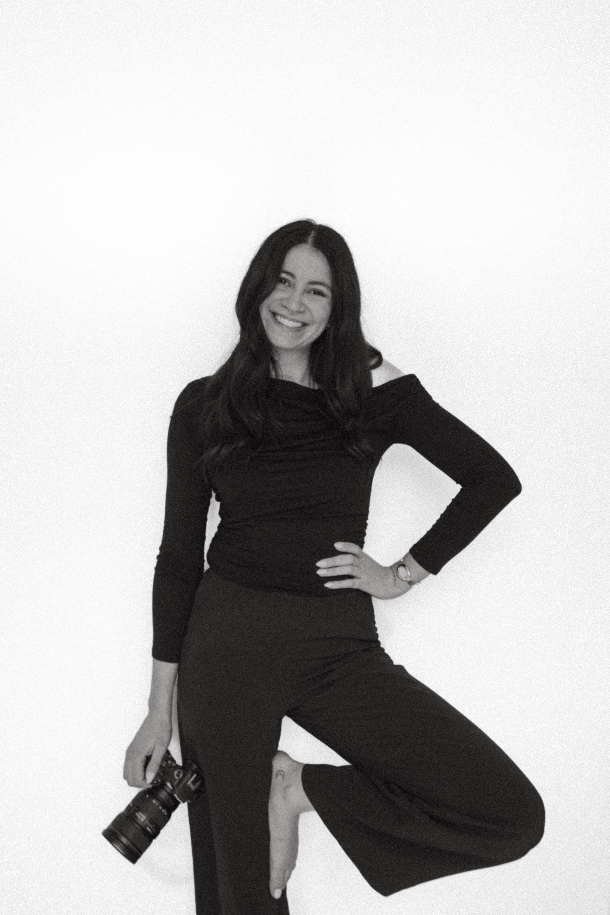 Black and white photo of a woman smiling, wearing a long-sleeved top, with a camera hanging from her right hand, sitting with one knee bent, against a plain white background.