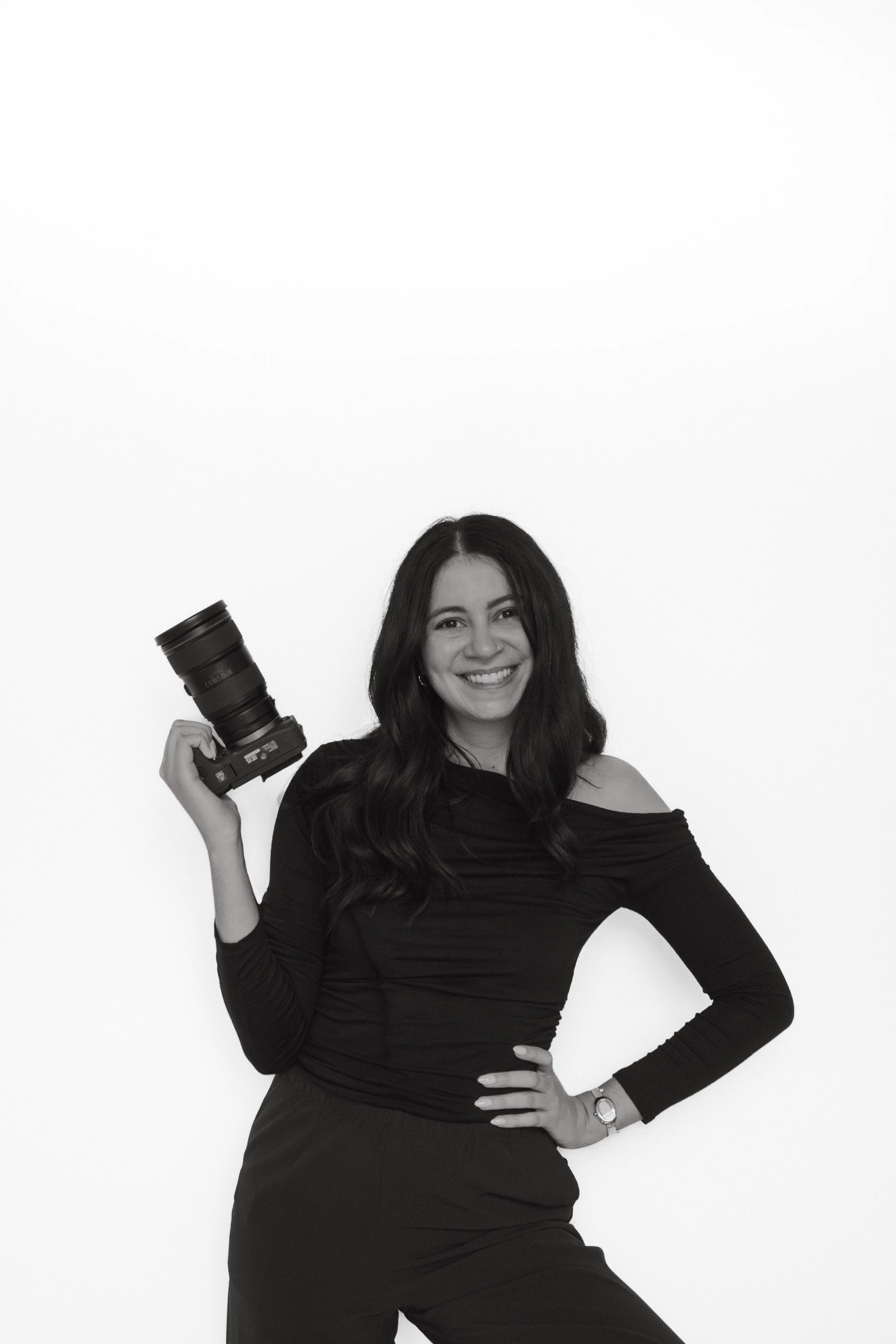 A woman wearing a long sleeve top and skirt, smiling, holding a camera with her right hand, and resting her left hand on her hip, against a plain white background.