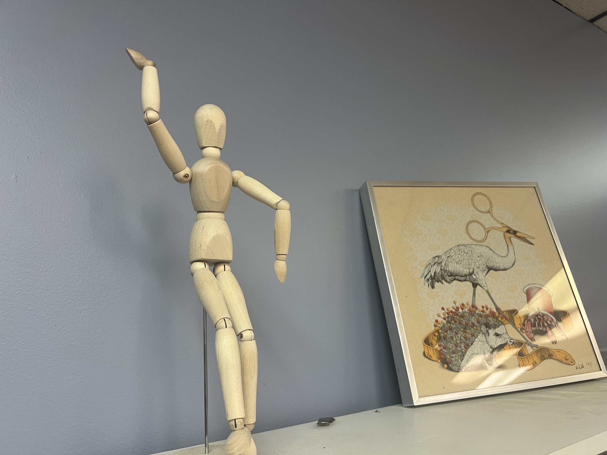 A wooden artist mannequin posed with its right arm raised above its head, against a light blue wall. To the right, there is framed artwork of a peacock with detailed feathers and a magnifying glass.