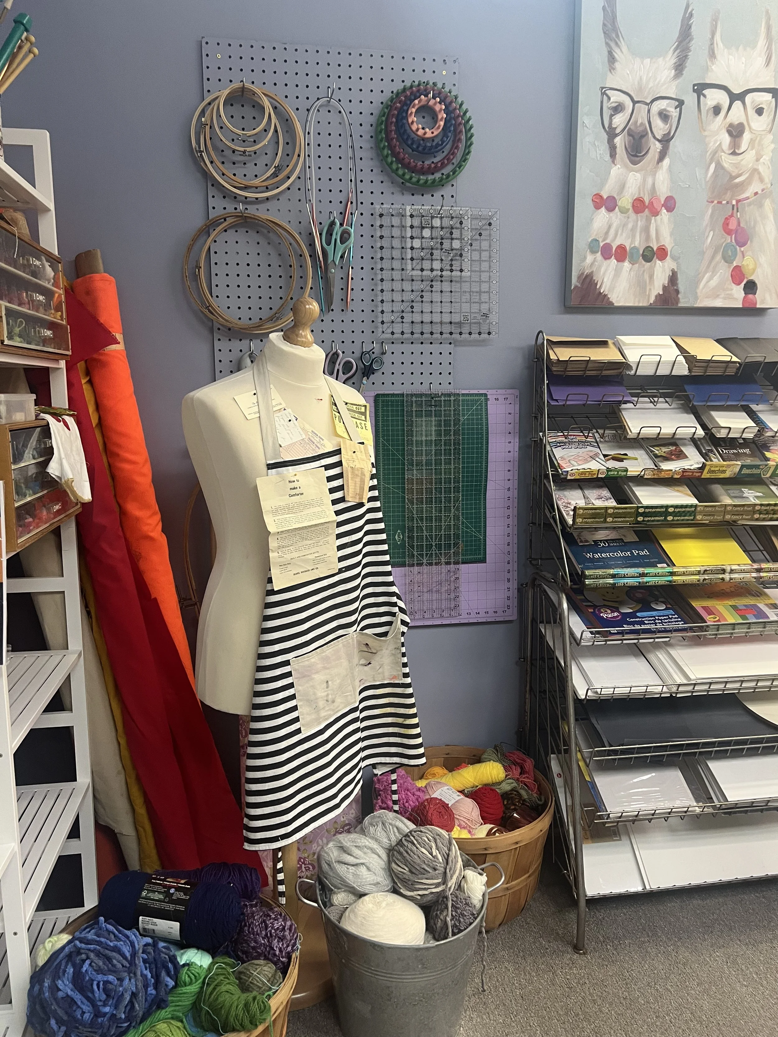A wall with a gray pegboard holding various embroidery hoops, scissors, and a measuring grid, with a colorful painting of two llamas wearing glasses and necklaces hanging on the wall. A dress form displays a striped apron, and there is a basket of yarn, along with shelves of craft supplies.