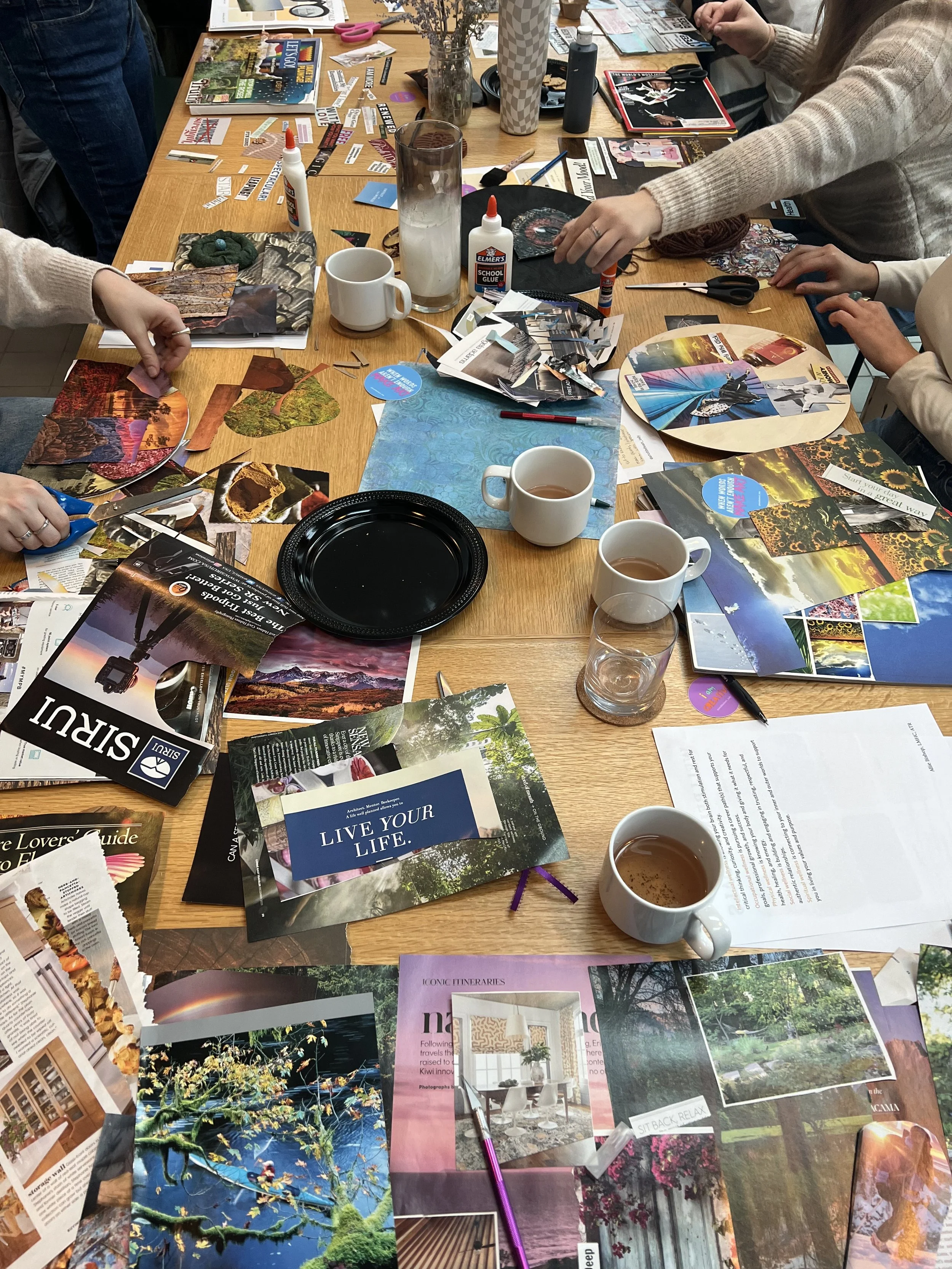 A cluttered wooden table covered with various magazines, photos, scissors, glue, pens, and cups of tea. Several hands are visible, some cutting pictures and arranging them. The magazines and photos feature nature scenes, landscapes, and interior design ideas.