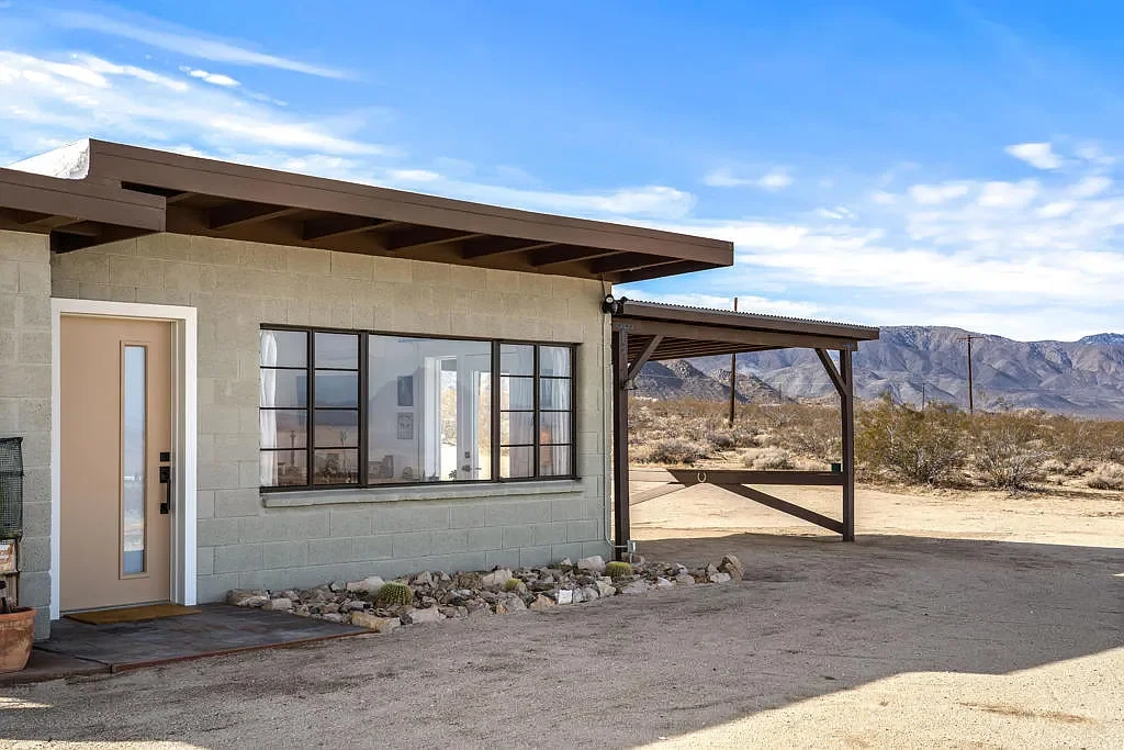 A small rural house with a beige front door and large window, set in a desert landscape with mountains in the background under a partly cloudy sky.