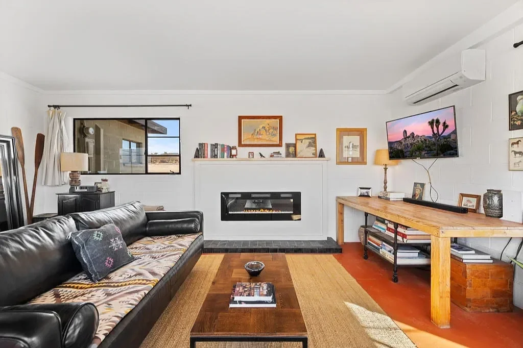 Living room with black leather sofa, wooden coffee table, fireplace, flat-screen TV on white brick wall, wooden console table, and framed photos and artwork.