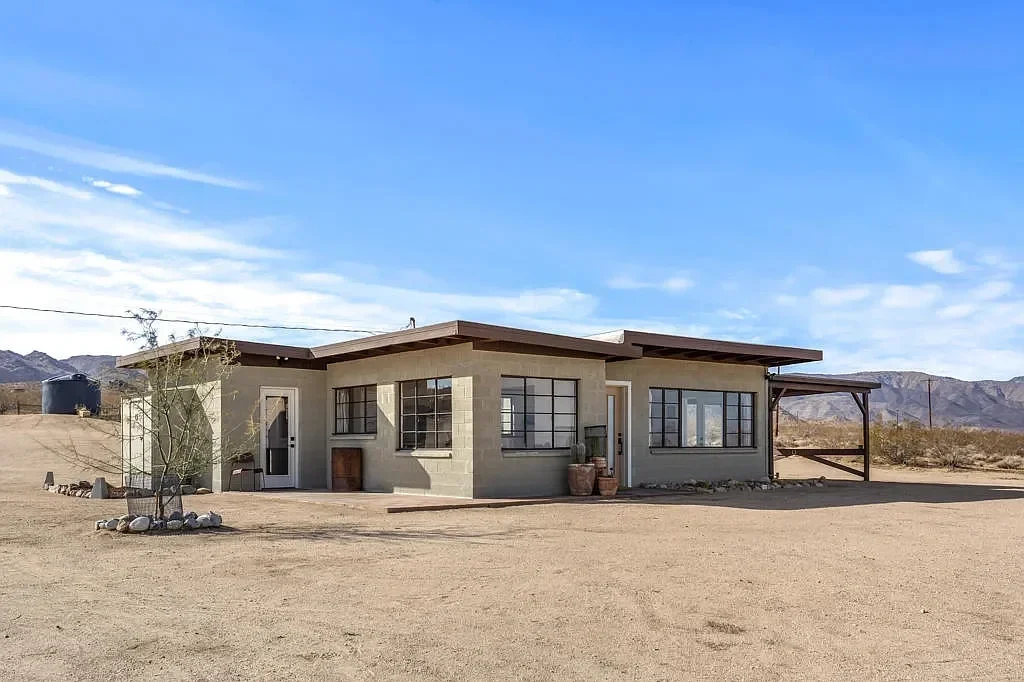 Single-story house with large windows, situated in a dry, desert landscape under a blue sky.