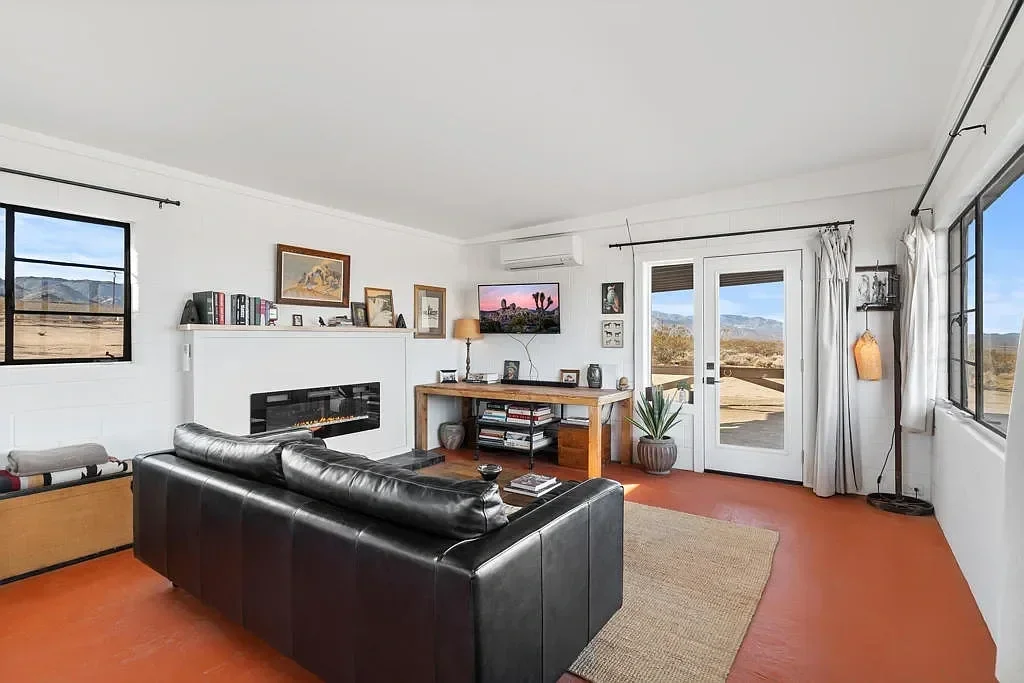 Living room with a black leather sofa, fire feature, bookshelf above, wooden console with decor and books, potted plant, wall-mounted TV, large windows, and glass door showing desert landscape outside.