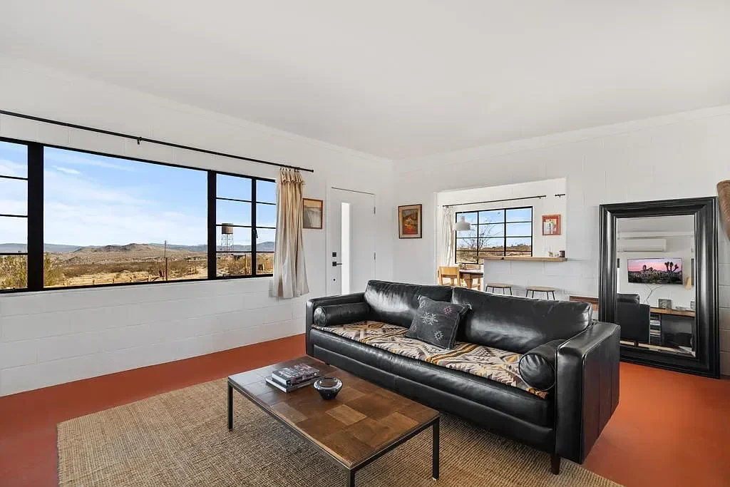Living room with large windows showing a desert landscape, black leather sofa, wooden coffee table, mirror, and small dining area.