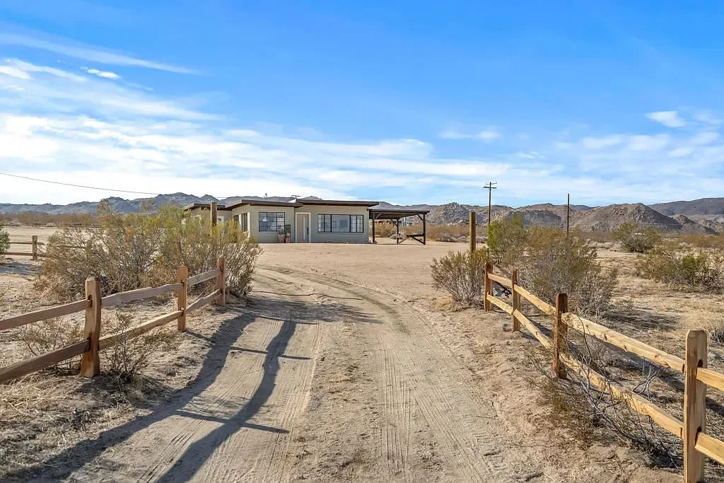 A desert landscape with a dirt driveway leading to a house, surrounded by sparse bushes and mountains in the background under a partly cloudy sky.