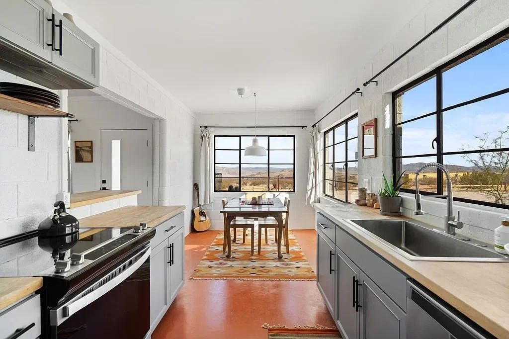 Bright kitchen with white brick walls, gray cabinets, wooden countertops, large black-framed windows, a dining table, tableware, a guitar, and a colorful rug.