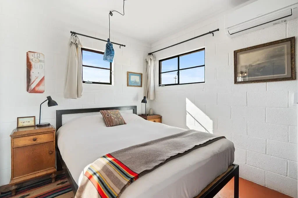 A bedroom with white brick walls, two windows with beige curtains, a bed with white bedding and a colorful striped blanket, and bedside tables with lamps. Artwork hangs on the walls.