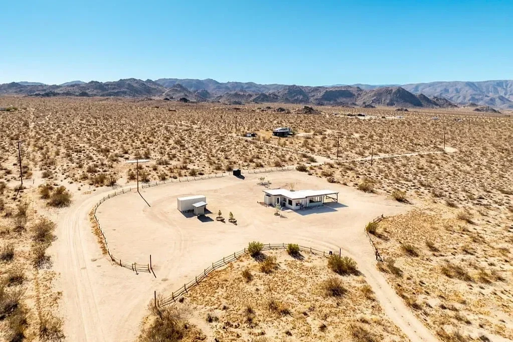 Aerial view of a small house and detached shed surrounded by desert landscape, with a gravel driveway, fences, and distant mountain range.