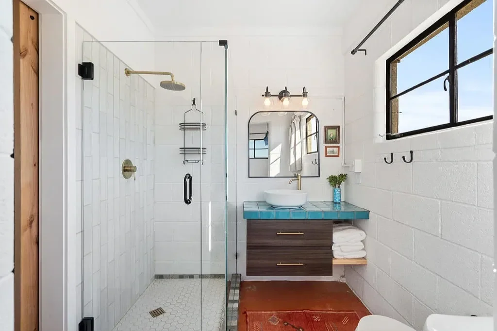 Modern bathroom with walk-in shower, white brick walls, blue tiled countertop with white vessel sink, large window, wall-mounted mirror, and mounted light fixtures.