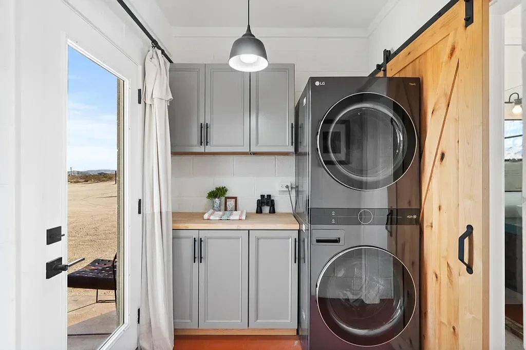Laundry room with stacked grey washing machine and dryer, grey cabinets, wood sliding barn door, and a glass door leading outside.
