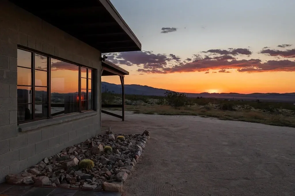 A sunset view from a house in a desert landscape, showing a rock garden with cacti outside a window with a mountain in the distance.