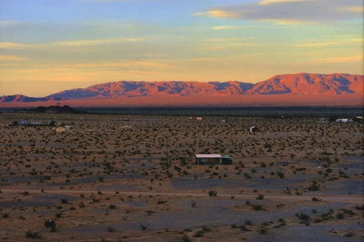 Desert landscape with sparse vegetation, small buildings, and a mountain range in the distance under a partly cloudy sky.