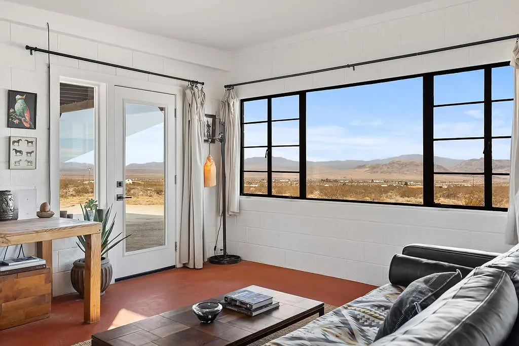 View of a living room with large windows showing a desert landscape with distant mountains, a white brick wall, a black leather chair, a wooden coffee table, and a door leading outside.