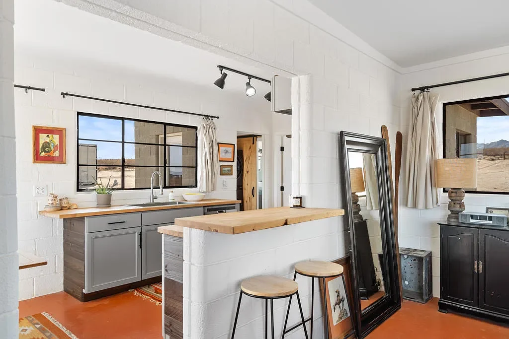 Bright kitchen with white walls, gray cabinets, wooden countertops, black window frame, and art on the walls. A mirror is leaning against the wall near a window with beige curtains. Decor includes a lamp, framed pictures, and stools.