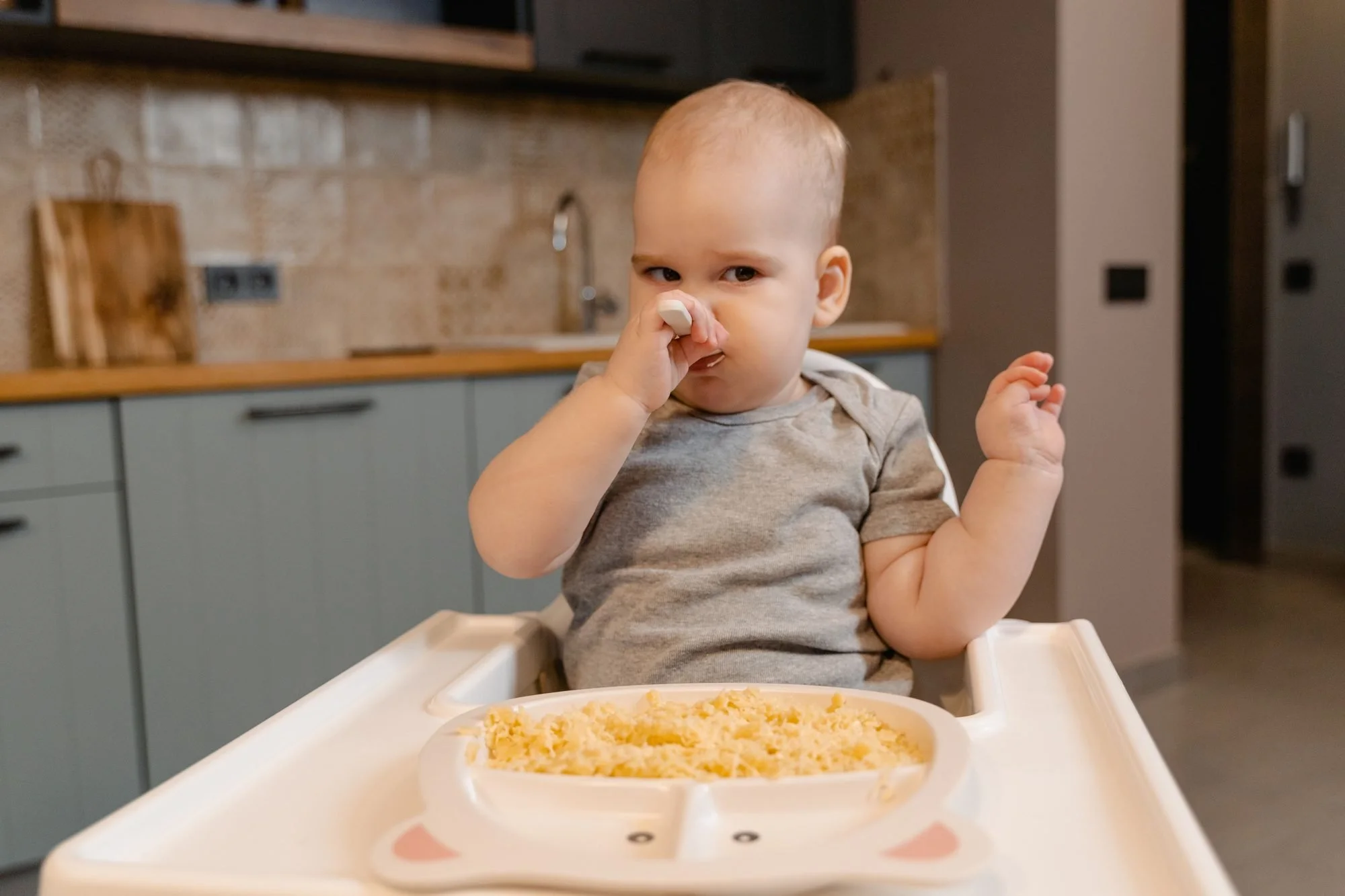 Baby in a white high chair holding a spoon to his mouth, with a divided plate of food on the tray, in a home kitchen.