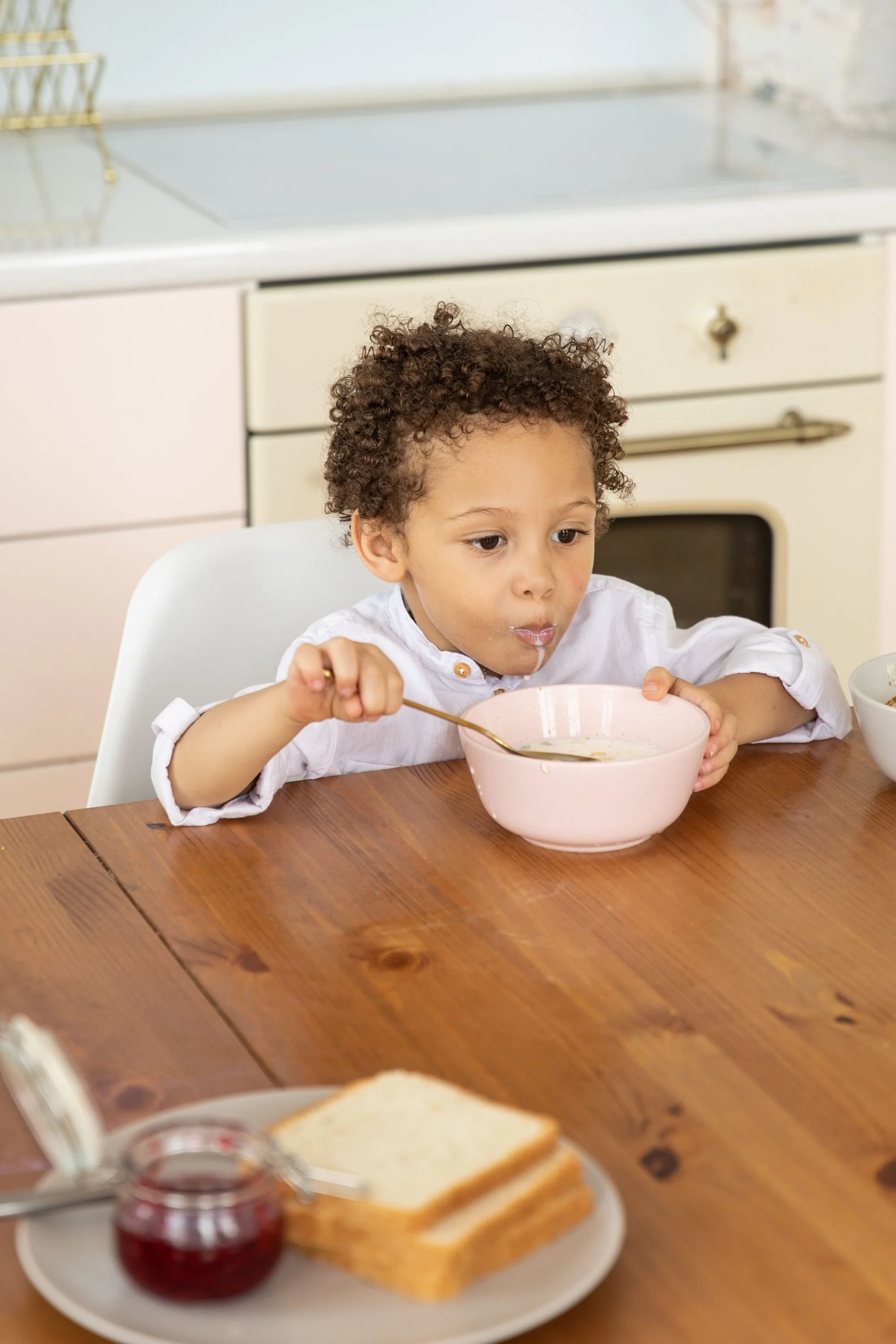 Young child with curly hair sitting at a wooden kitchen table, eating cereal from a pink bowl with a spoon, toast and jam in the foreground.