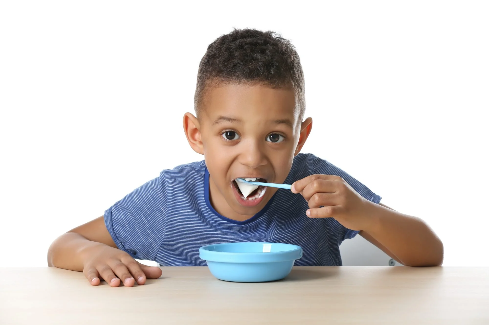 Young boy in a blue shirt sitting at a light wooden table, eating white food from a blue bowl with a blue spoon against a white background.