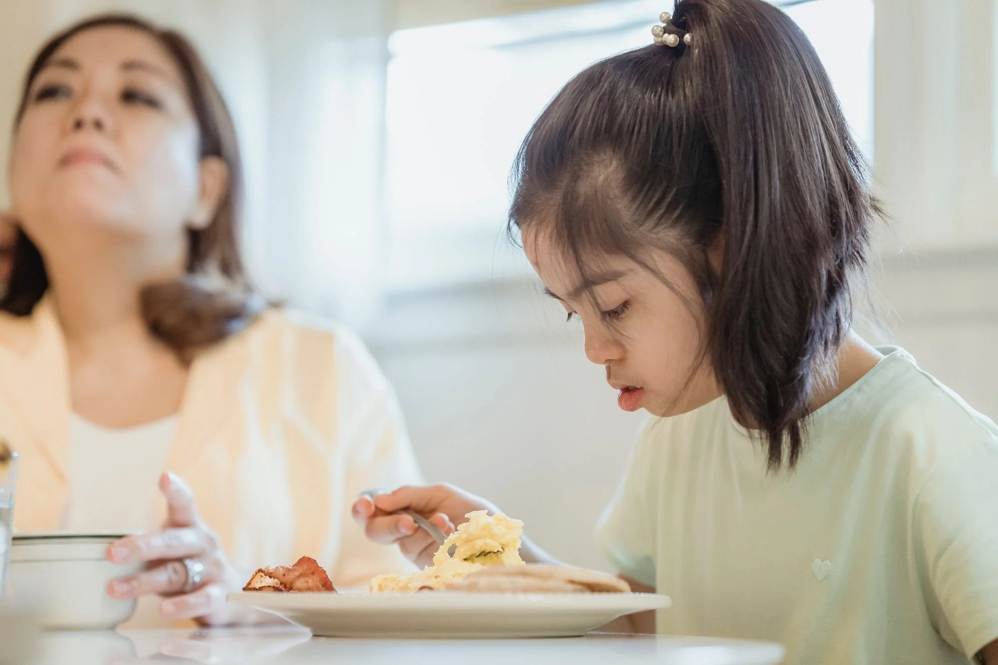 Young girl with Down syndrome eating scrambled eggs with a fork at a white table, with her mother sitting beside her in a bright kitchen.