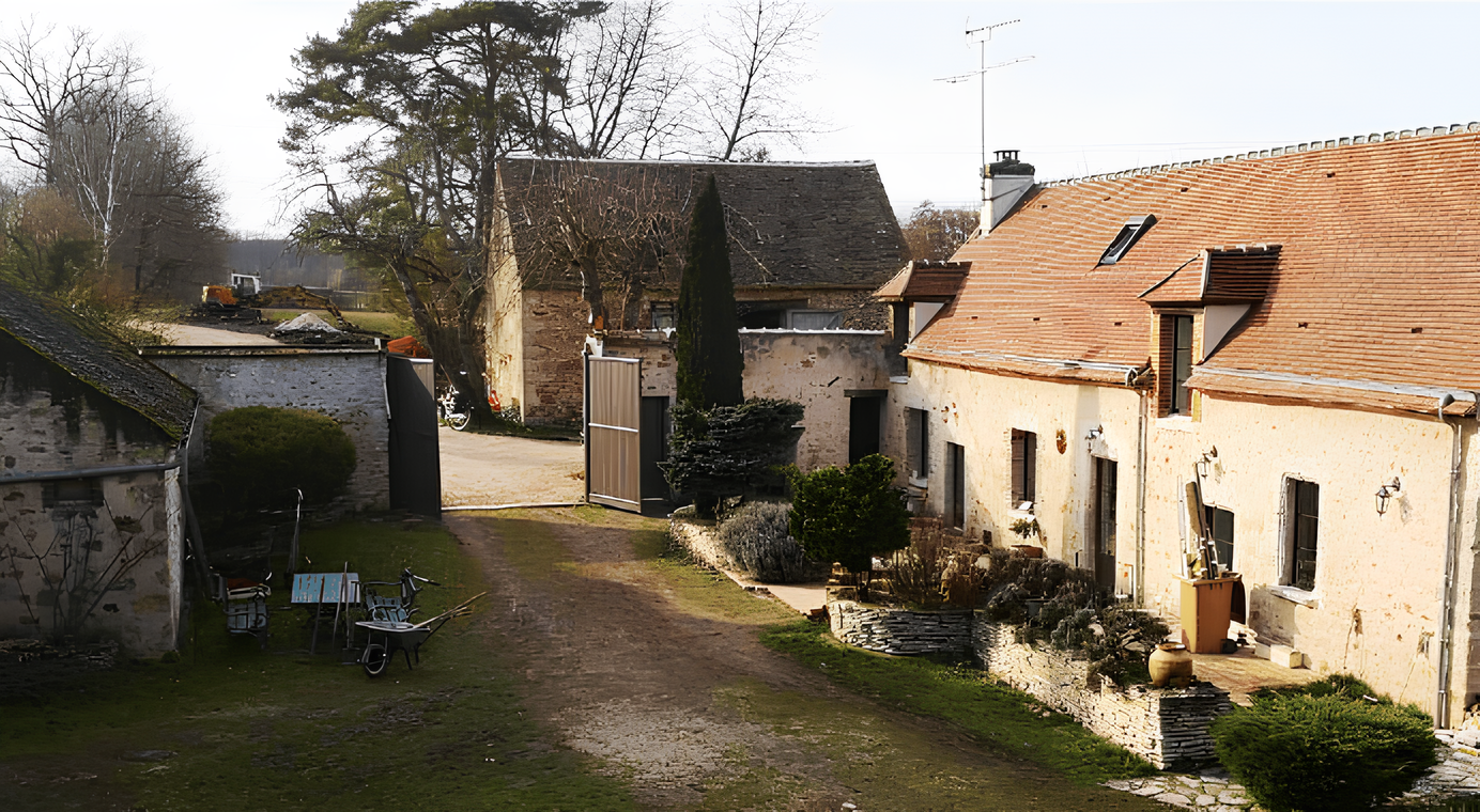 Une cour intérieure d'une ferme ou d'une maison ancienne avec des bâtiments en pierre et une allée en terre, quelques meubles de jardin et des arbres sans feuilles.