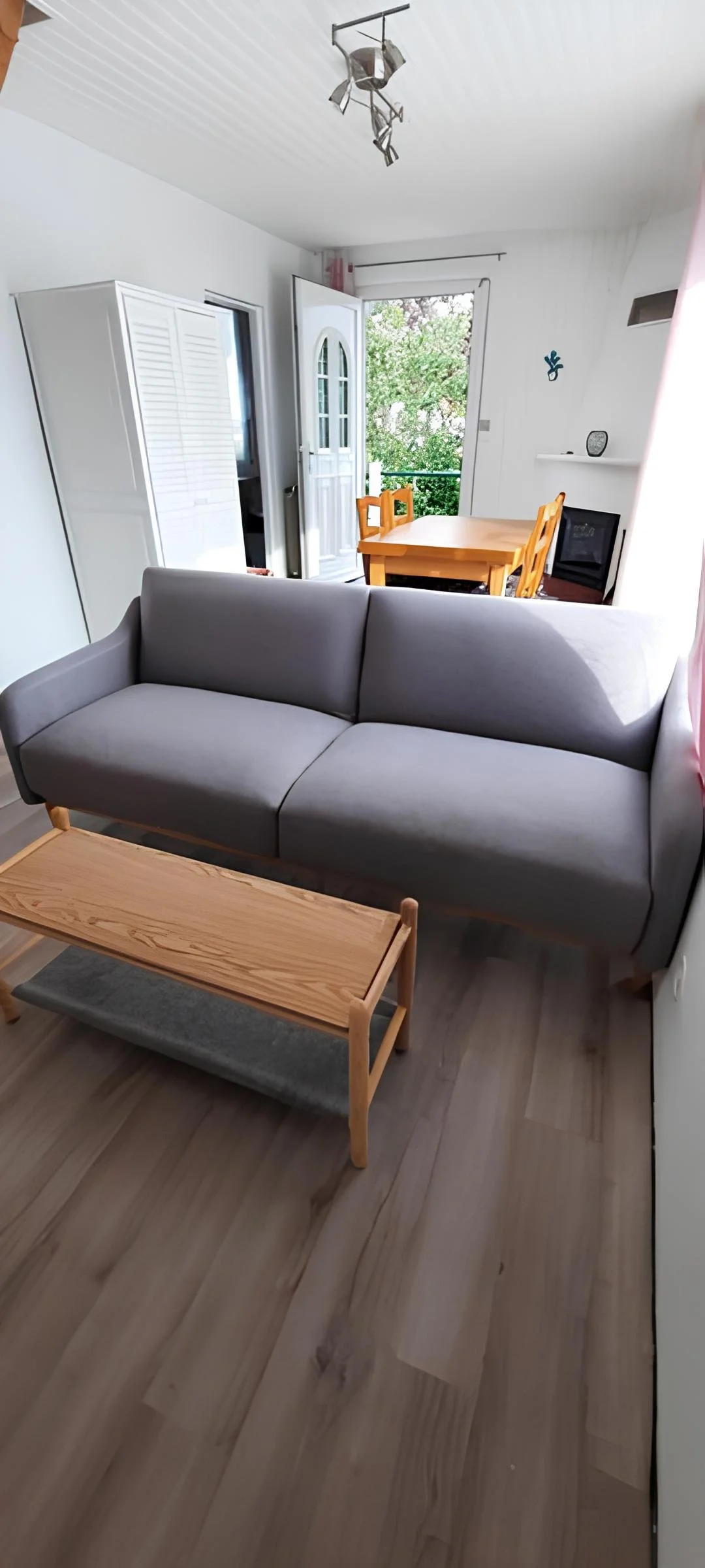 Un salon avec canapé gris, table basse en bois et porte donnant sur un balcon avec des chaises en bois, vue vers une salle à manger et un balcon en arrière-plan.