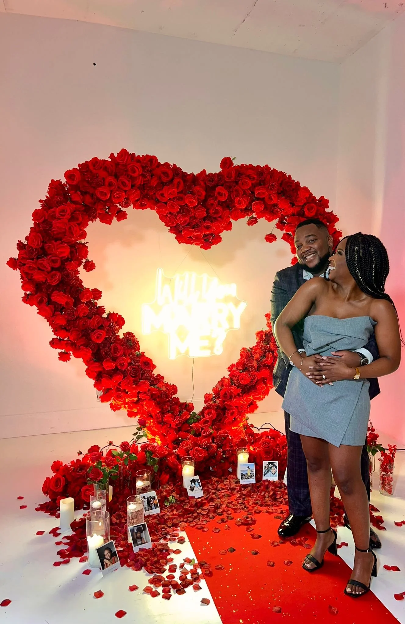 Un couple souriant, en costume, se tient devant un grand cœur en roses rouges, avec des bougies, des photos et des pétales de rose par terre. Il y a un panneau lumineux au fond qui dit : "J'aime MARRY ME ?" en forme de carte.