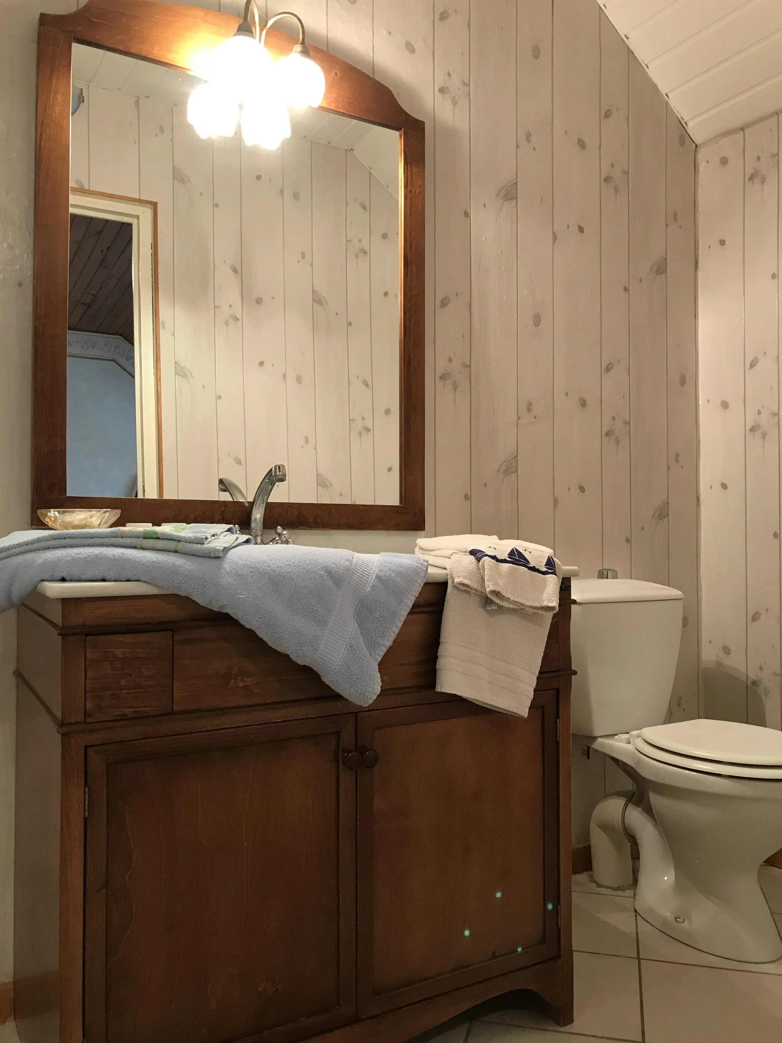Une salle de bain avec un miroir en bois, un lavabo avec des serviettes pliées dessus, un robinet, une cuvette de toilettes, et un mur recouvert de planches en bois clair.
